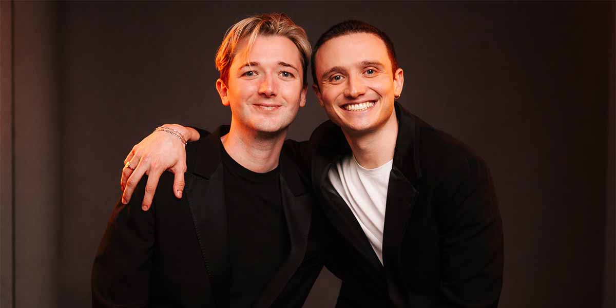 Two men pose closely, smiling. One wears a black jacket, the other a white shirt.