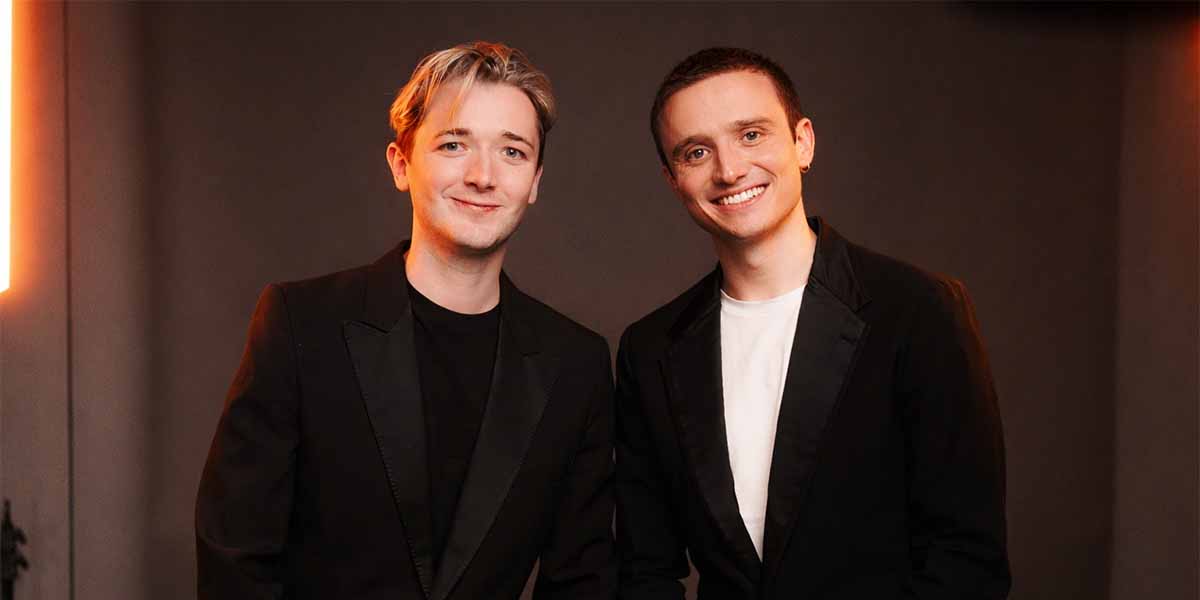 Two men in black blazers smile at the camera against a neutral background with soft lighting.