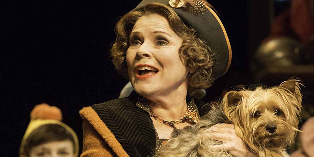 A woman with curly hair, wearing a hat, smiles while holding a small dog on stage.