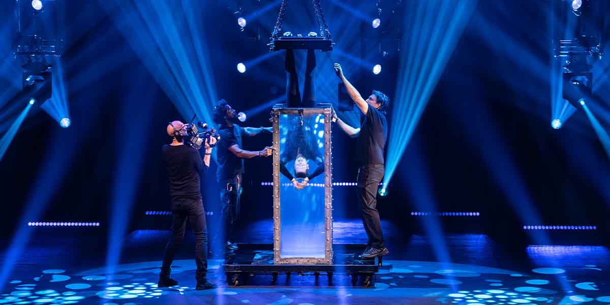 Andrew Basso suspended in a glass box, surrounded by performers and dramatic stage lighting.