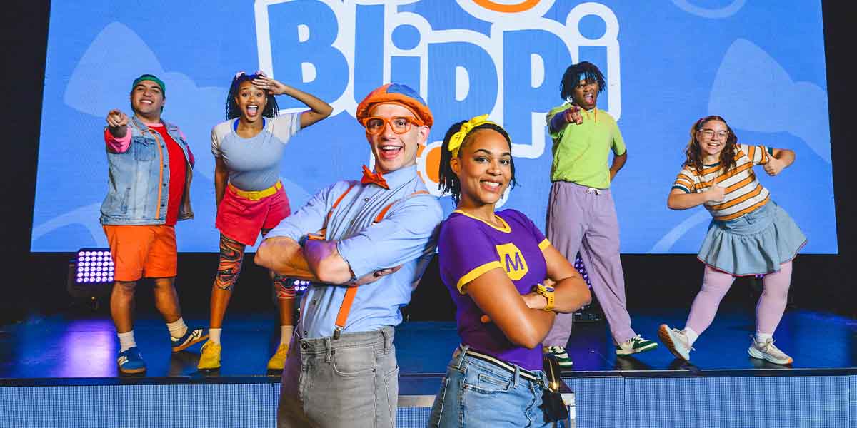 Six performers pose playfully on stage, wearing colourful outfits, with a large "Blippi" logo in the background.