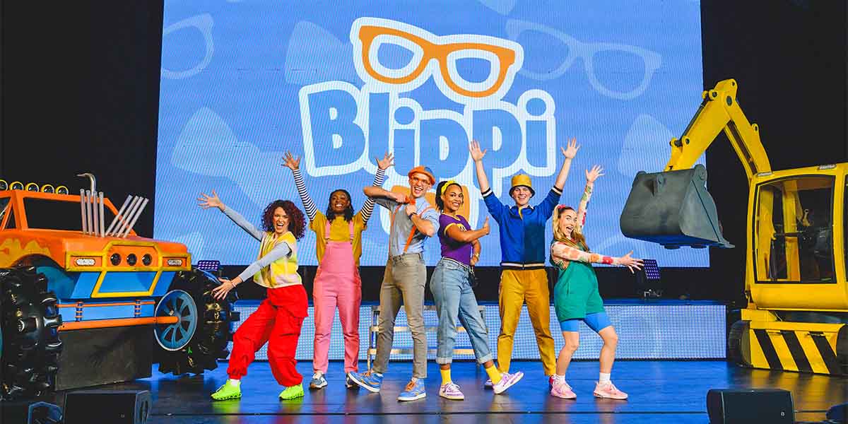 Seven performers in colourful costumes pose energetically on stage, with a large "Blippi" backdrop and machinery.