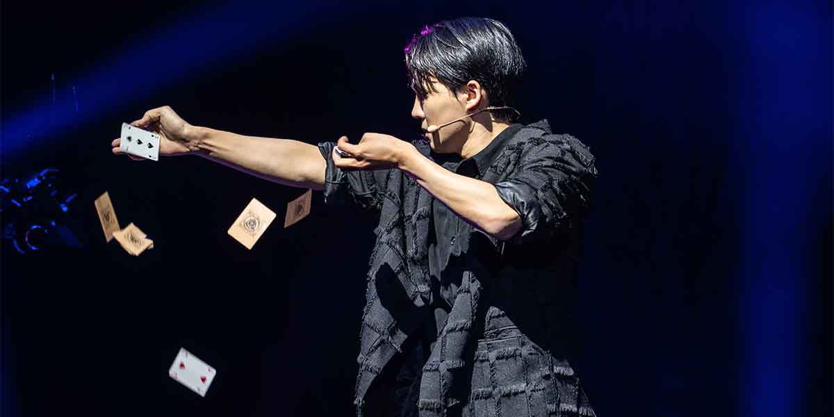 Eden Choi performs with playing cards in hand, wearing a textured black outfit against a dark background.