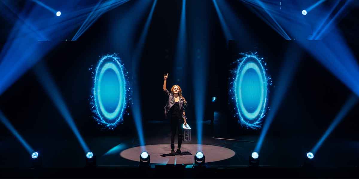 Gabriella Lester stands on stage, raising one hand, surrounded by blue light effects and spotlights.