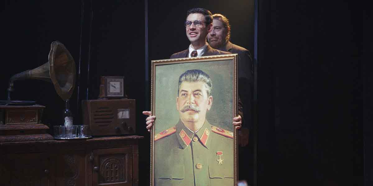 A man in a suit holds a portrait of Stalin, with a gramophone and vintage radio in the background.