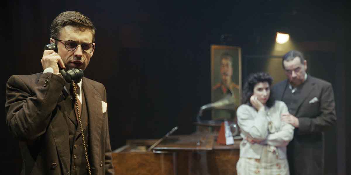 A man in glasses speaks on the phone, while a woman and another man stand nearby, concerned.