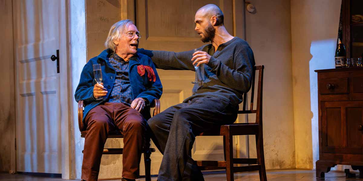 Jeff Rawle and Jack Riddiford sit together, sharing a moment, with a glass in hand, on stage.