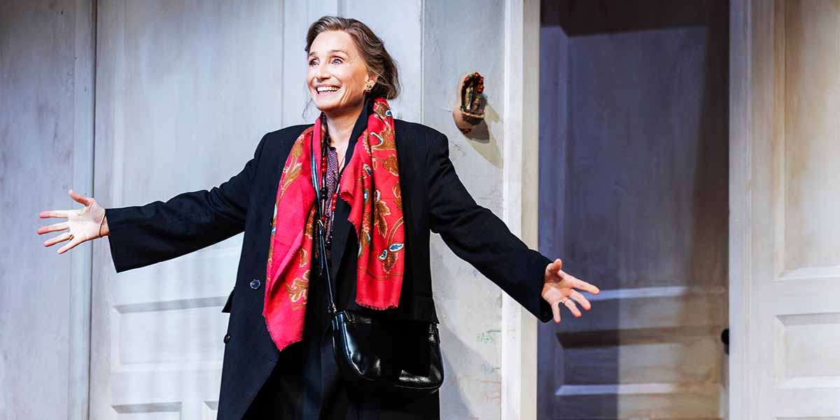 Kristin Scott Thomas smiles broadly, wearing a black coat and red scarf, with arms outstretched.