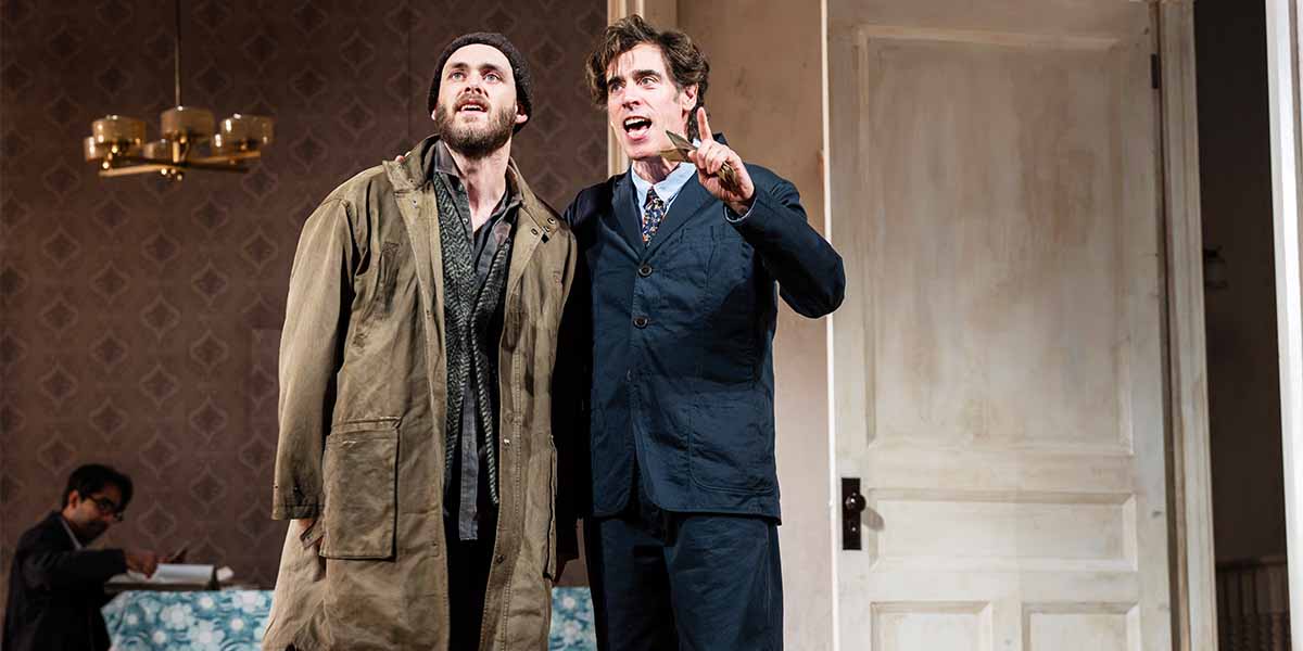 Jack Riddiford and Stephen Mangan stand together, gesturing expressively, in a theatrical setting with a door behind.