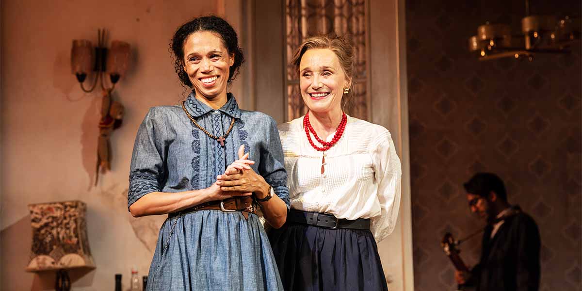Vinette Robinson and Kristin Scott Thomas smile together on stage, dressed in period costumes, with a musician in background.