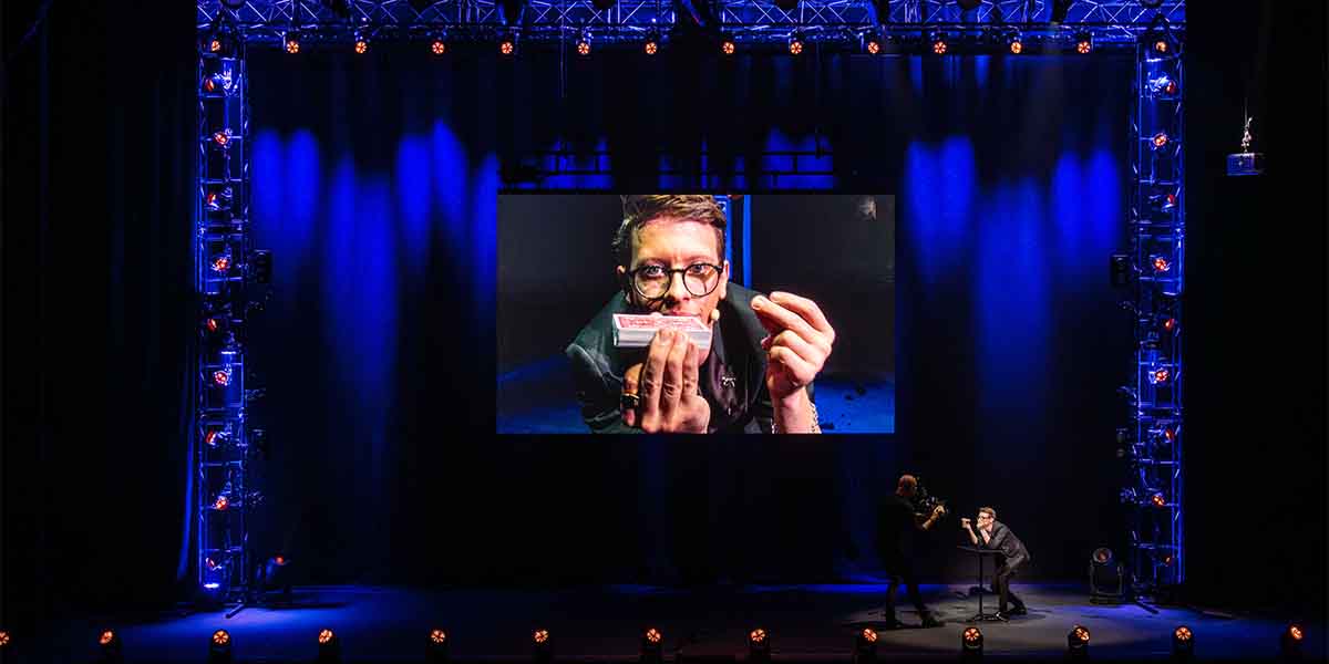 Matthew Pomeroy performs on stage, holding a card, with a large screen displaying his actions.
