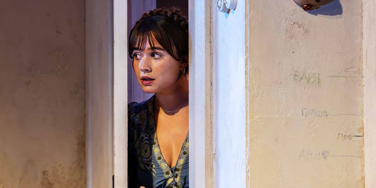 Megan Cusack peeks through a doorway, wearing a patterned dress, against a textured wall background.