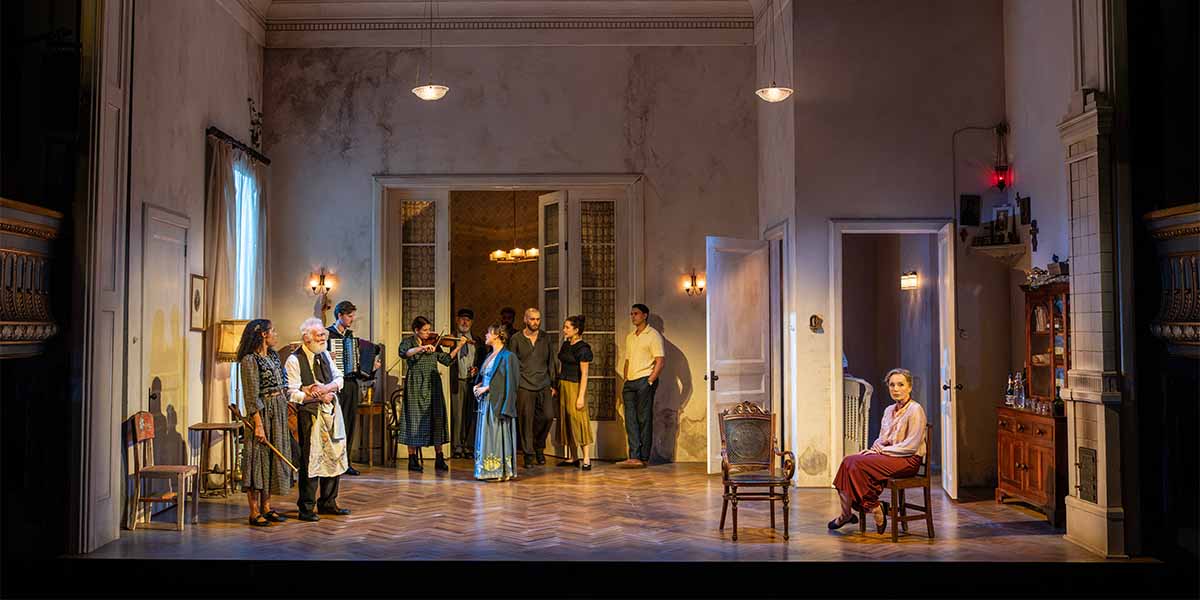 A group of actors in period costumes stands on stage, with one woman seated, in a domestic setting.
