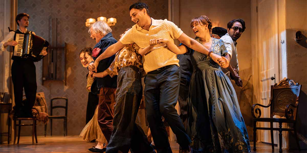 A group of actors joyfully dance together on stage, wearing period costumes, with an accordionist in the background.