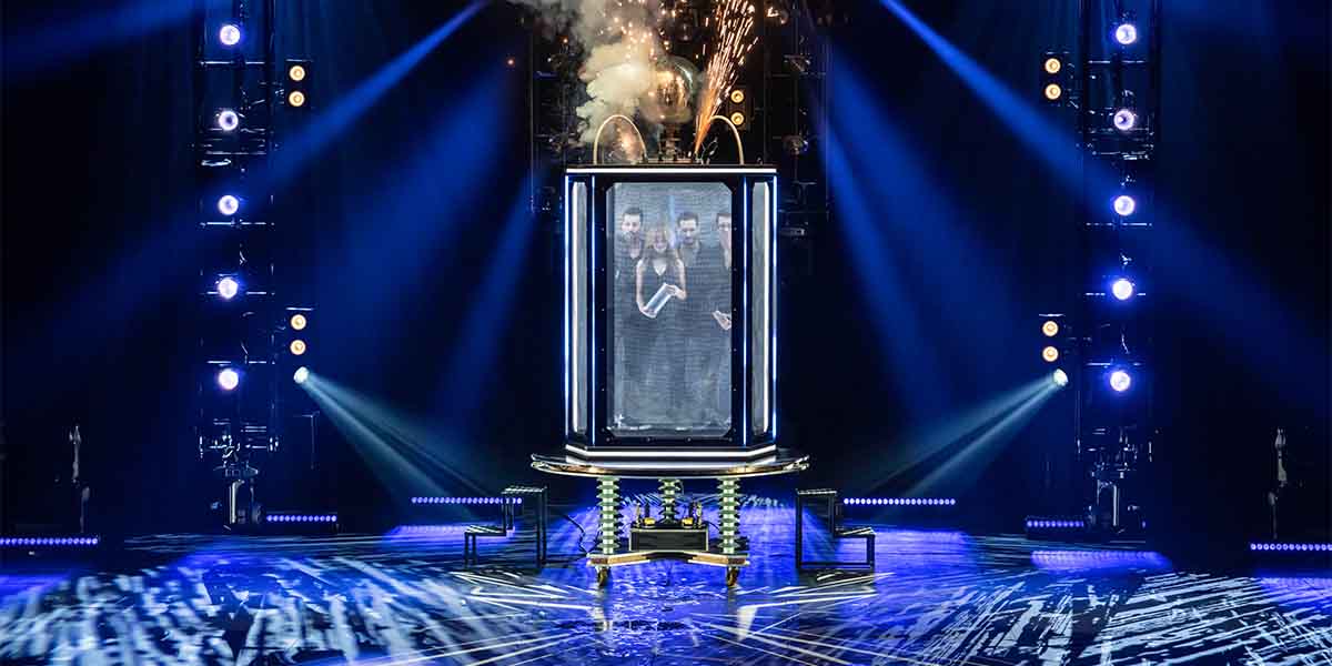 The Horsemen perform inside a glass box, surrounded by dramatic lighting and smoke effects on stage.