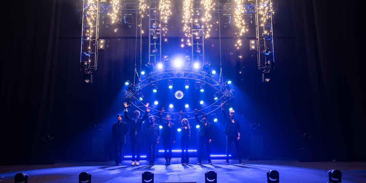 The cast of NOW YOU SEE ME LIVE stands on stage, illuminated by blue lights and sparkling effects.