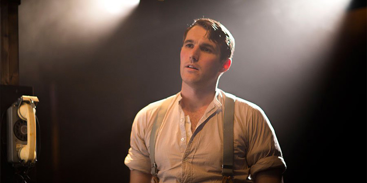 George Turvey stands under dramatic lighting, wearing a white shirt and suspenders, with a vintage telephone nearby.