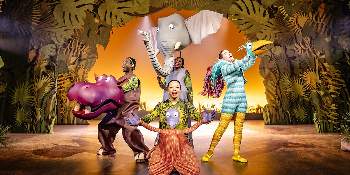 A crocodile puppet in a vibrant costume, surrounded by children in a lively theatre setting.