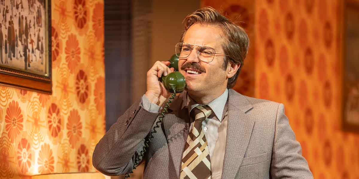 A man with glasses and a moustache smiles while speaking on a green telephone in a retro setting.
