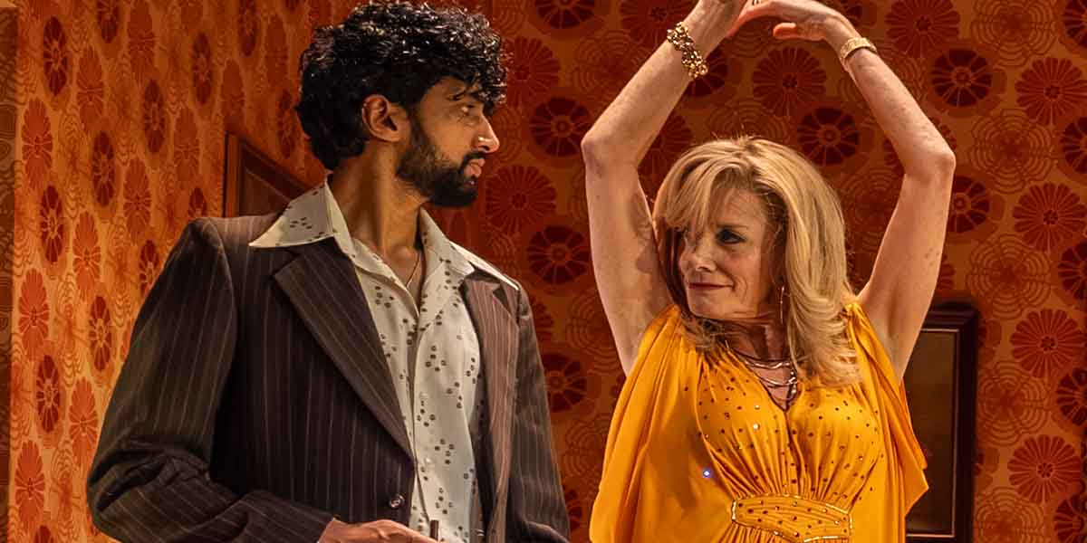 A man in a striped suit watches a woman in a yellow dress dance with raised arms.