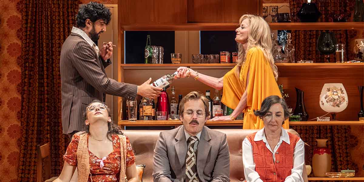 A man hands a bottle to a woman in an orange dress, while three others observe.