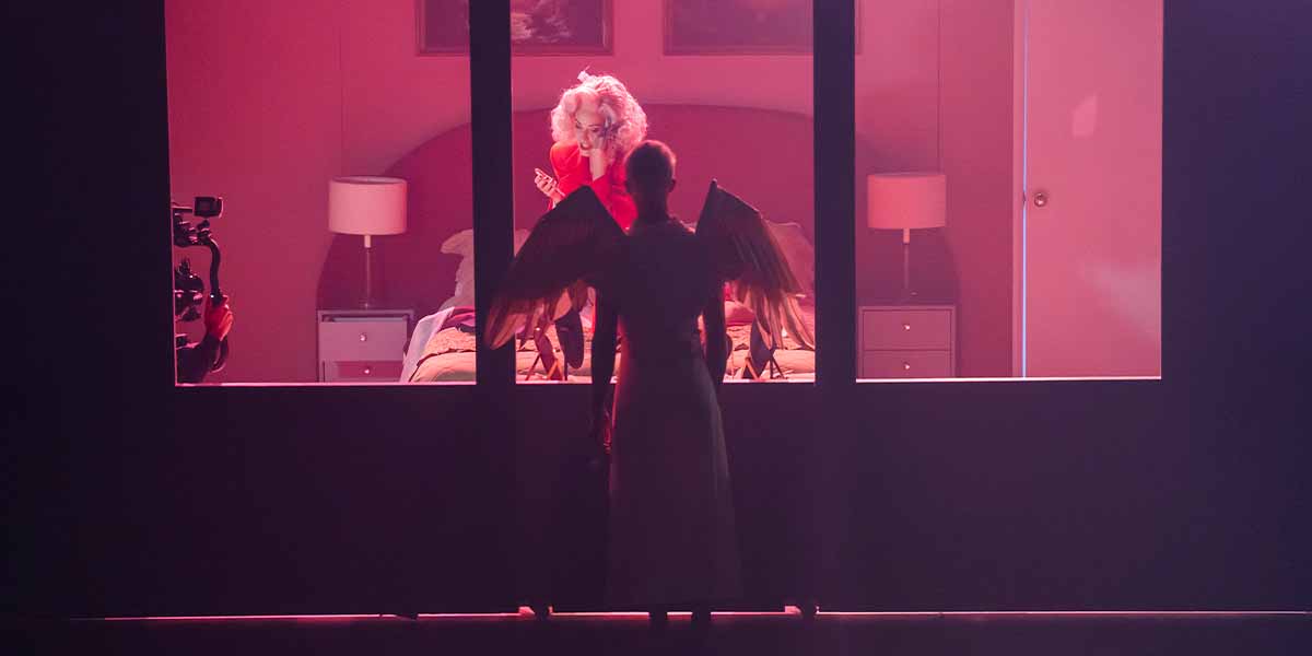 Allison Cook in red, with a figure in white and wings, framed by a pink-lit window.