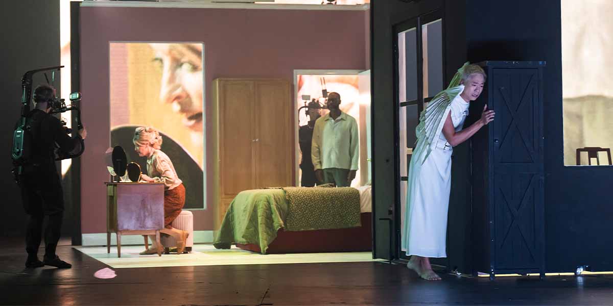 Allison Cook sits at a vanity, while a character with wings peeks from behind a door.