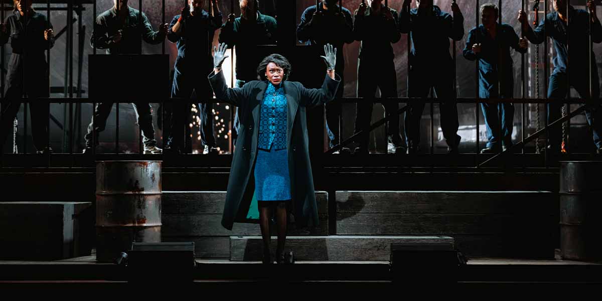Cassiopeia Berkeley-Agyepong stands centre stage in a blue dress, raising her hands, surrounded by ensemble members.