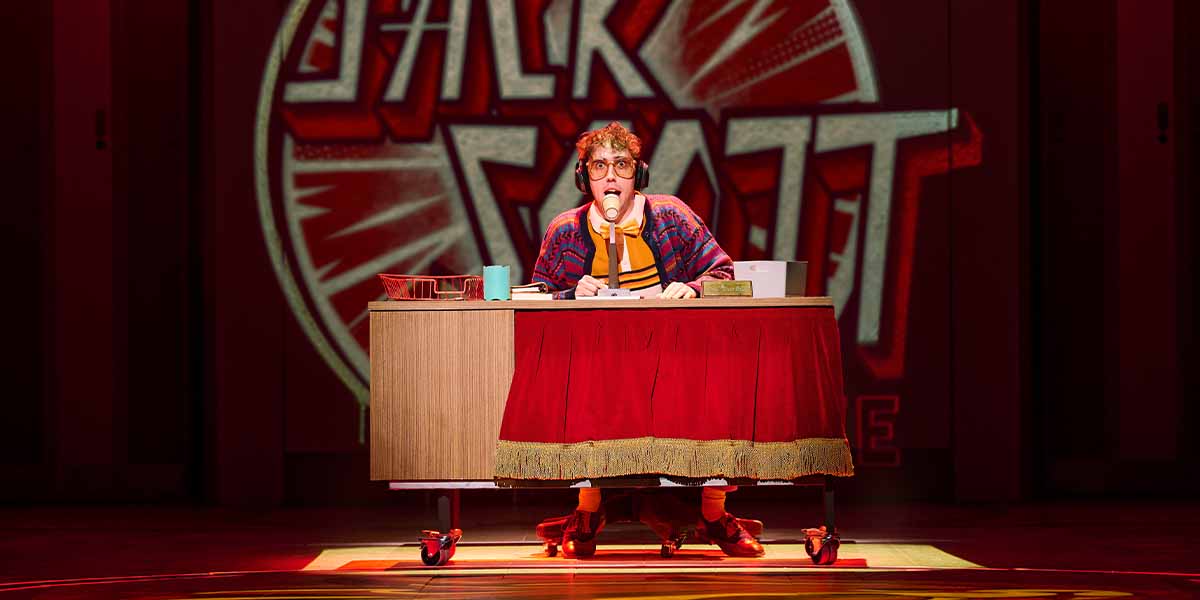 Curtis Patrick as Jack Scott sits at a desk, wearing headphones and holding a microphone, with vibrant background.
