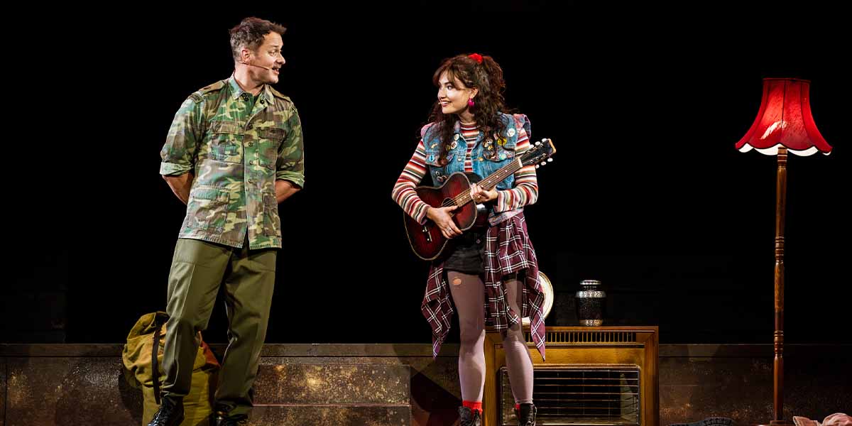 Declan Bennett in military attire and Hannah Richardson with a guitar perform on stage, with a lamp nearby.
