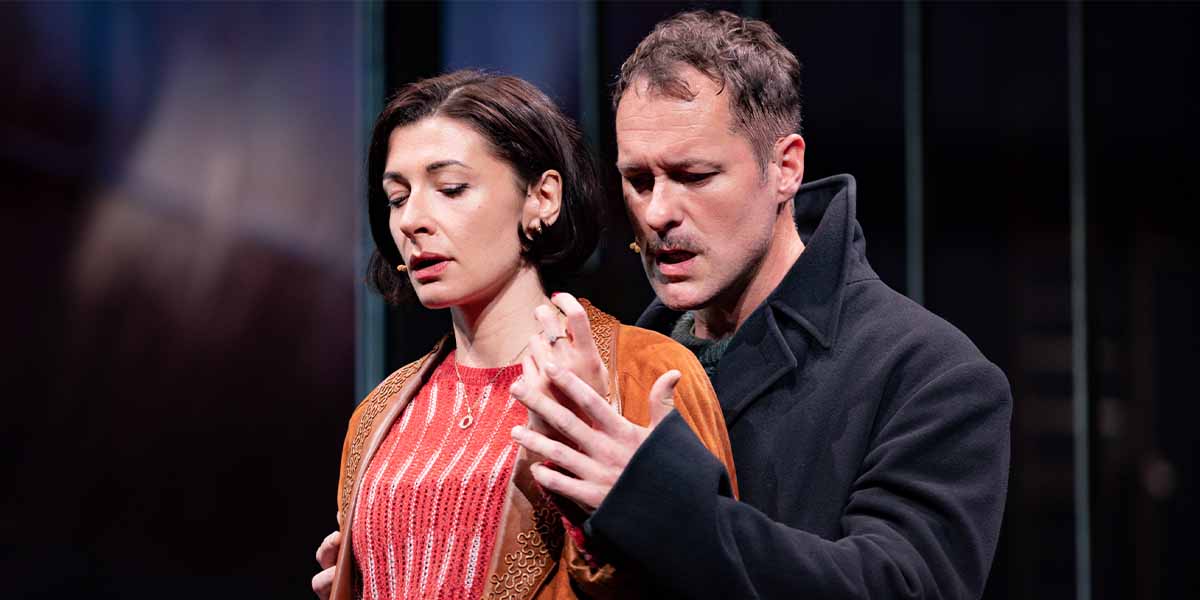 Frances McNamee and Declan Bennett perform closely together, expressing emotion, against a dark, blurred background.