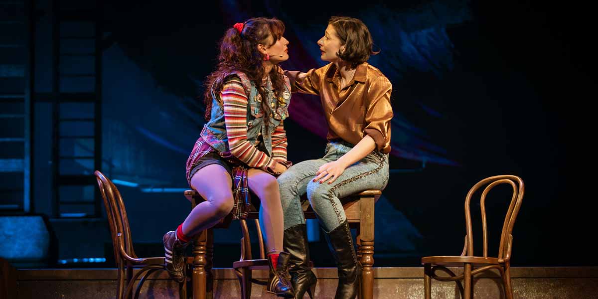 Hannah Richardson and Frances McNamee sit closely on stools, engaged in conversation, against a dark backdrop.