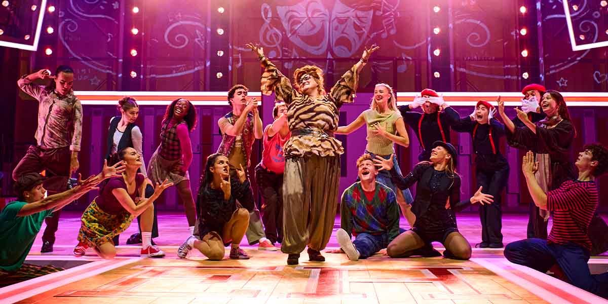 KayCee Stroh as Ms Darbus stands centre stage with the cast of High School Musical, vibrant costumes visible.