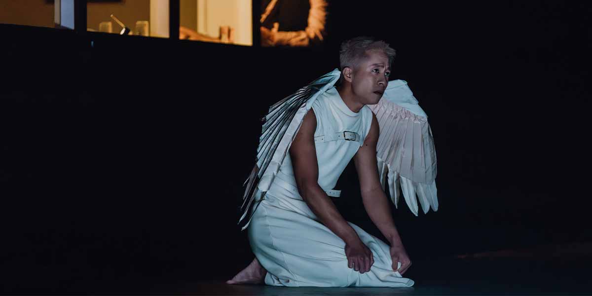 Keith Pun, dressed in a white costume with wings, kneels on stage, expressing emotion against a dark background.