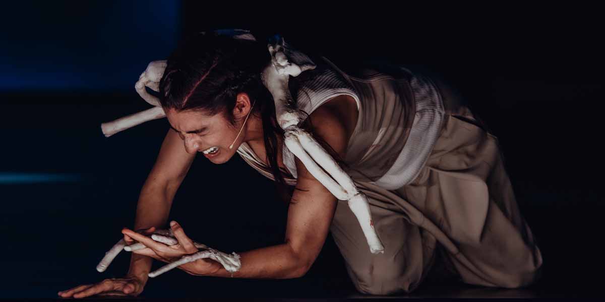 Mariam Wallentin performs on stage, kneeling with expressive anguish, wearing a flowing beige costume and skeletal props.