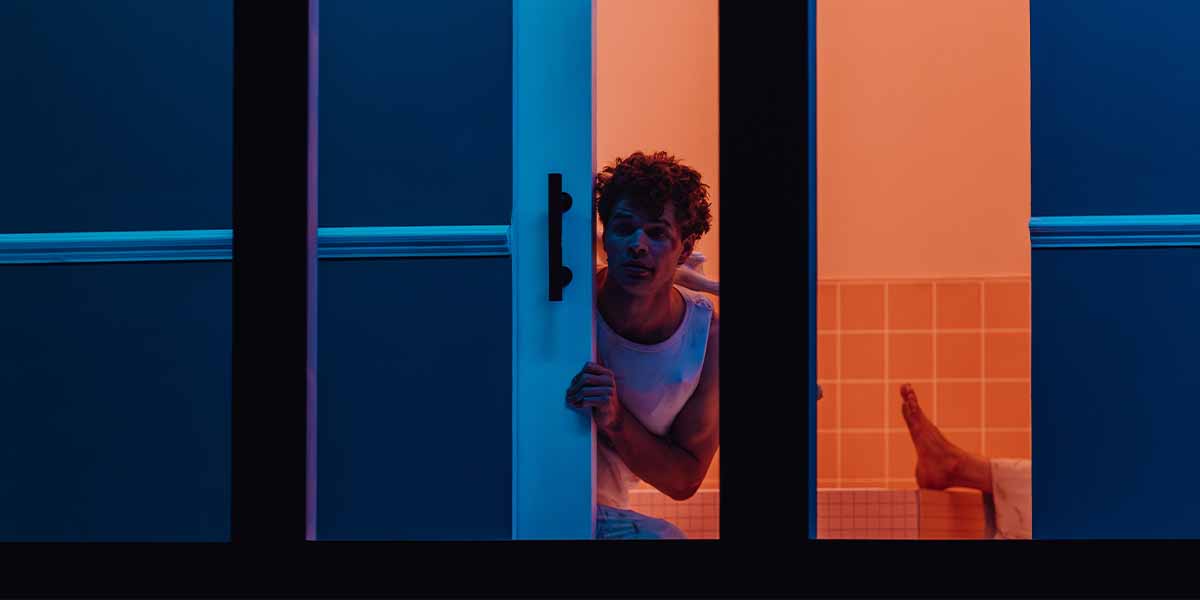 Matthew McKinney peers through a partially open door, with warm orange and cool blue lighting contrasting.