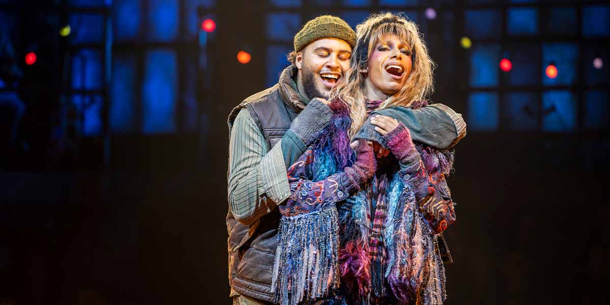 Billy Nevers as Collins and Jeevan Braich as Angel embrace joyfully, wearing vibrant, textured costumes.
