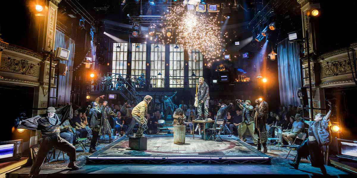 The full cast of RENT poses together, wearing vibrant costumes, with a colourful backdrop and stage lights.