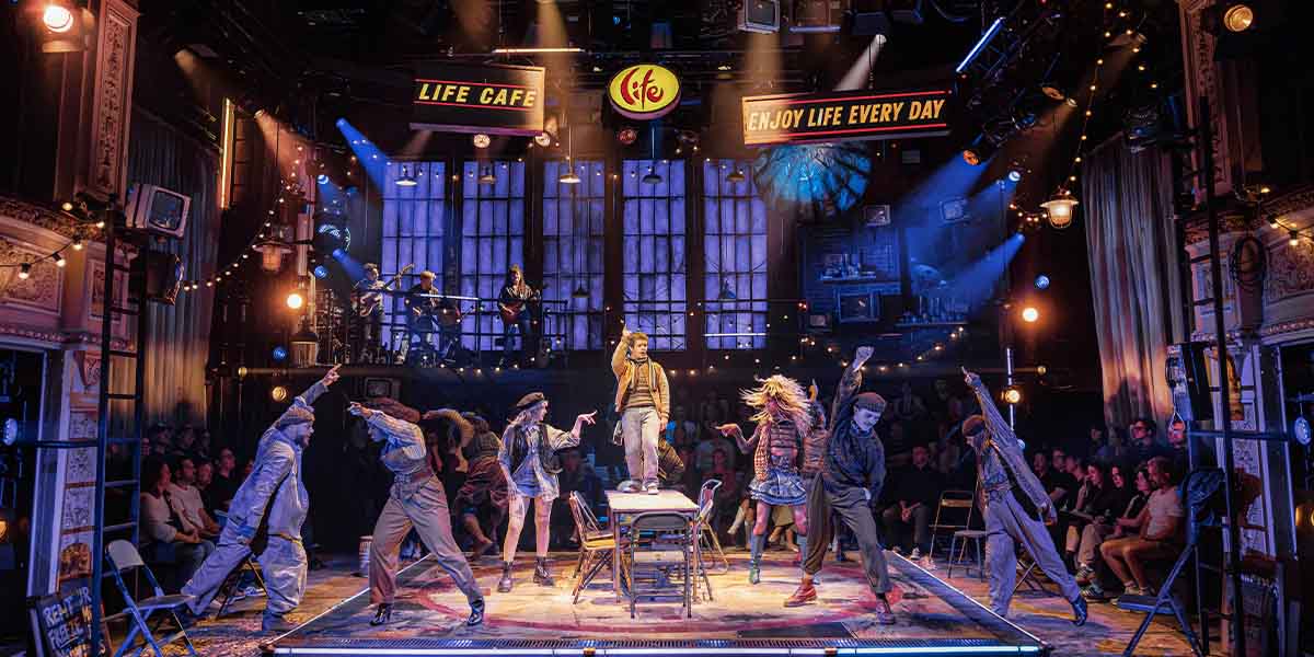 Gaten Matarazzo as Mark stands centre stage with the cast of RENT, vibrant costumes and dramatic lighting.