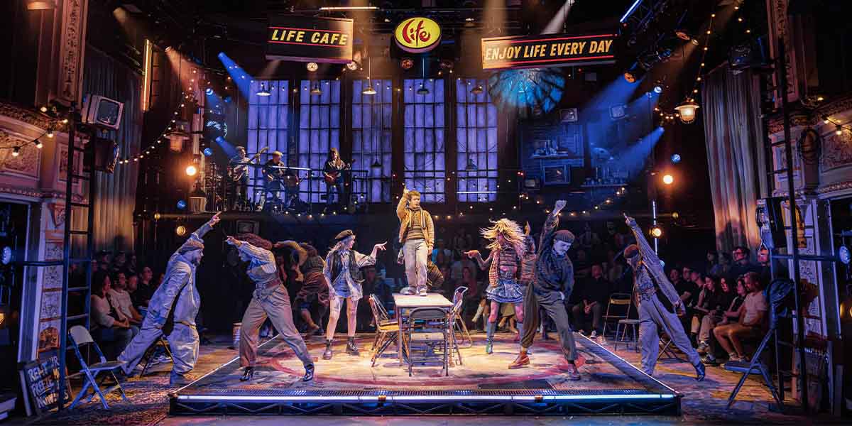 Gaten Matarazzo as Mark stands centre stage, surrounded by cast members in vibrant costumes, under dramatic lighting.