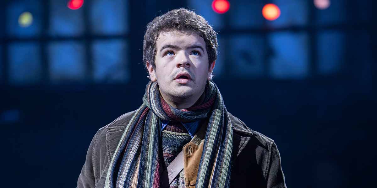 Gaten Matarazzo as Mark, looking upwards, wearing a brown coat and multi-coloured scarf, stage lights in background.