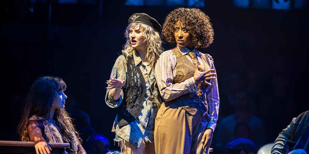 Maureen and Joanne perform on stage, expressing emotions; audience members are blurred in the background.
