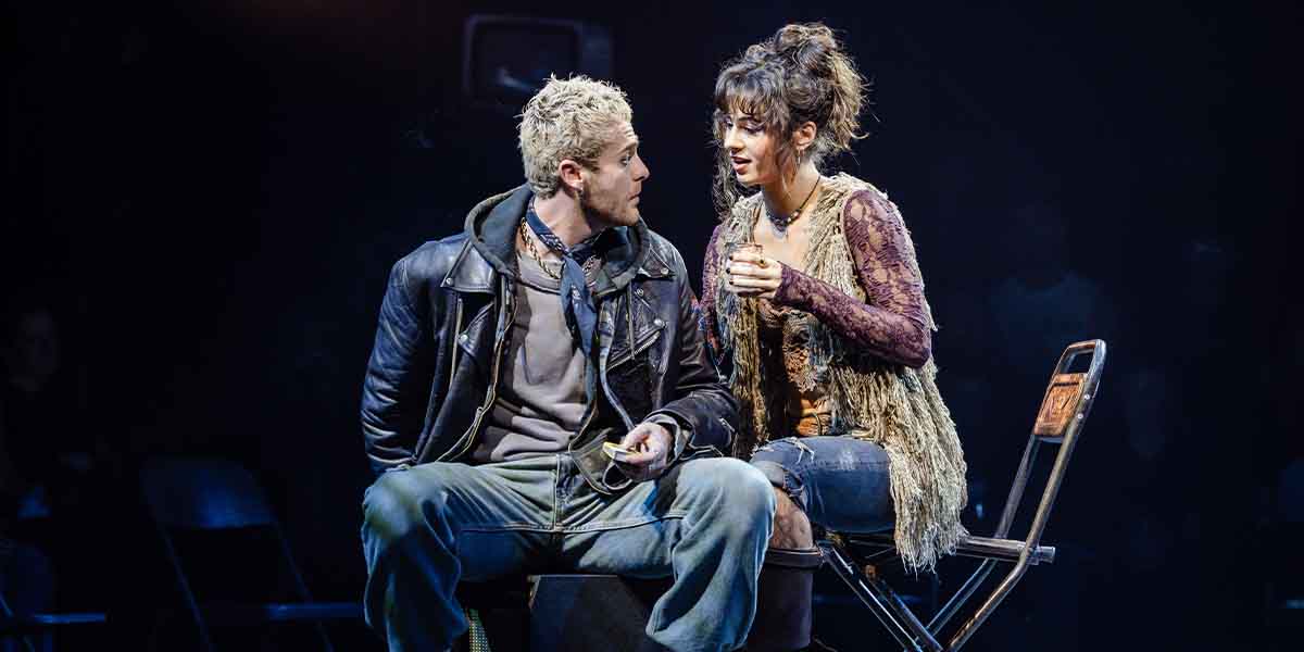 Travis Ross as Roger and Alexa Lopez as Mimi sit closely, engaged in conversation, under dramatic lighting.