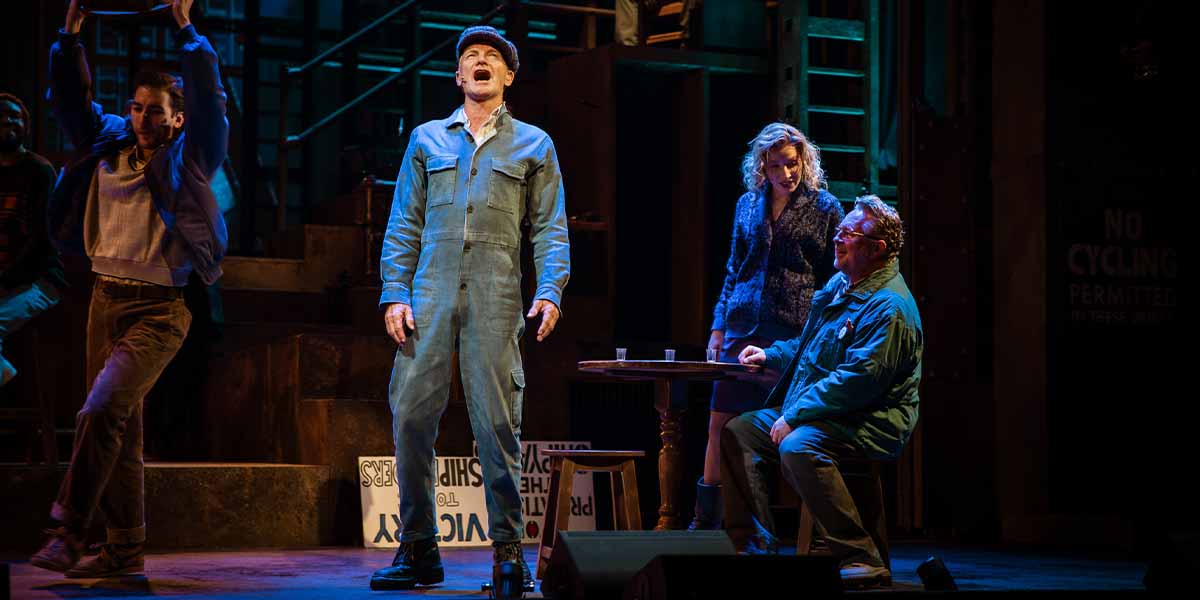 Sting performs centre stage in a blue jumpsuit, surrounded by three other actors in a dramatic scene.
