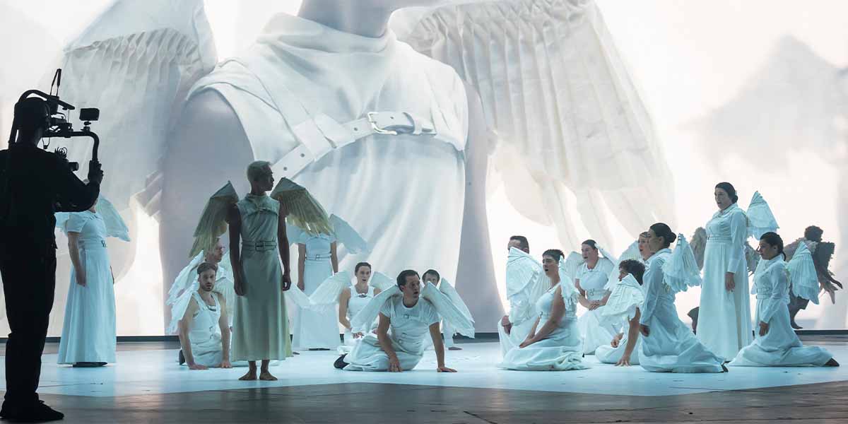 A group of performers in white costumes with wings, staged against a large, ethereal backdrop.