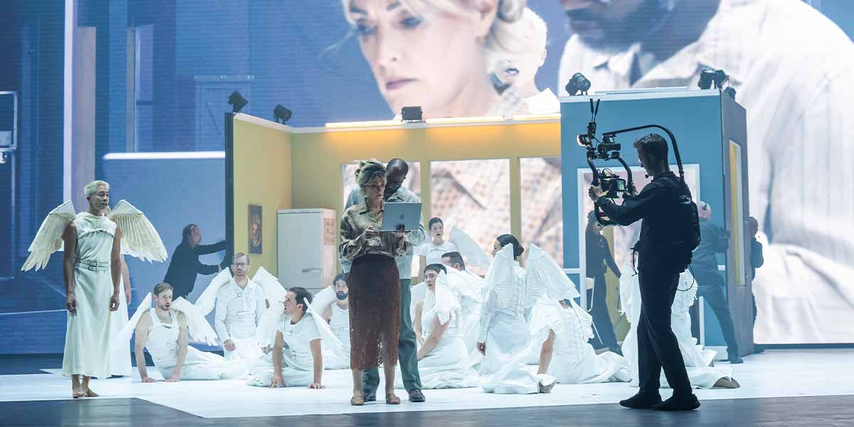 A diverse cast performs on stage, featuring angels in white and a woman with a laptop.