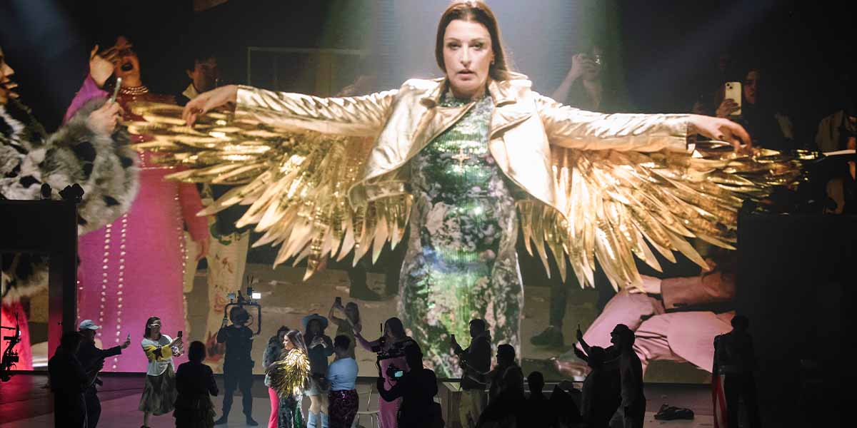 A performer in a gold winged jacket stands centre stage, surrounded by a lively cast and audience.