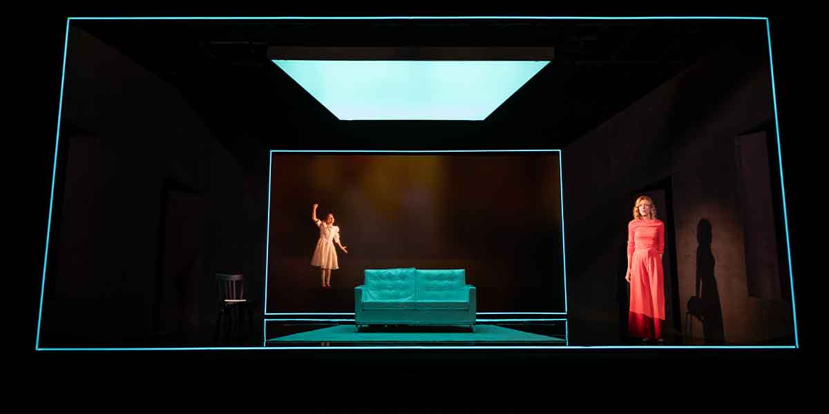 A person in a red dress stands beside a teal sofa, while another person in white appears to float.