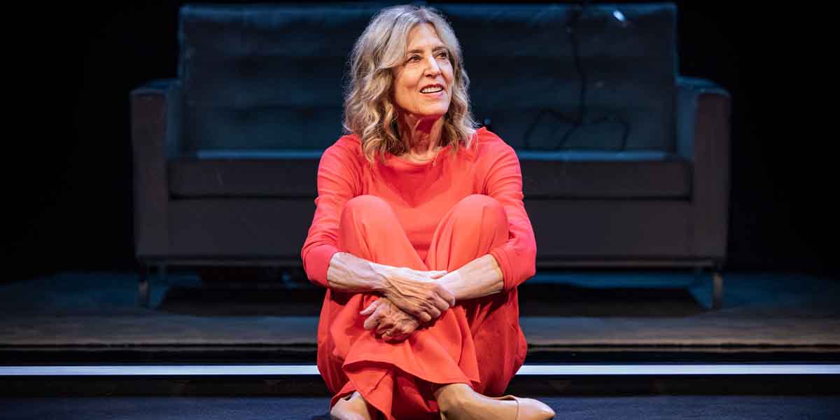 A person in a vibrant red outfit sits on the stage, smiling, with a black sofa in the background.