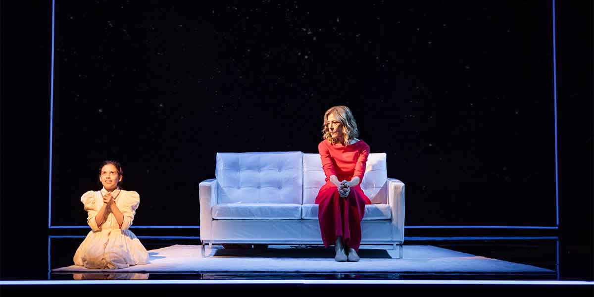 A person in a red dress sits on a white sofa, while a child in a white dress kneels beside.